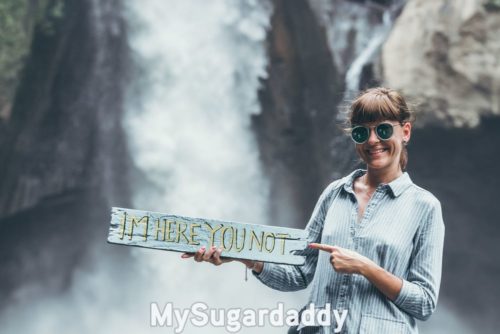 girl in front of waterfall holding board meant to cause envy that reads "I'm here, you not"