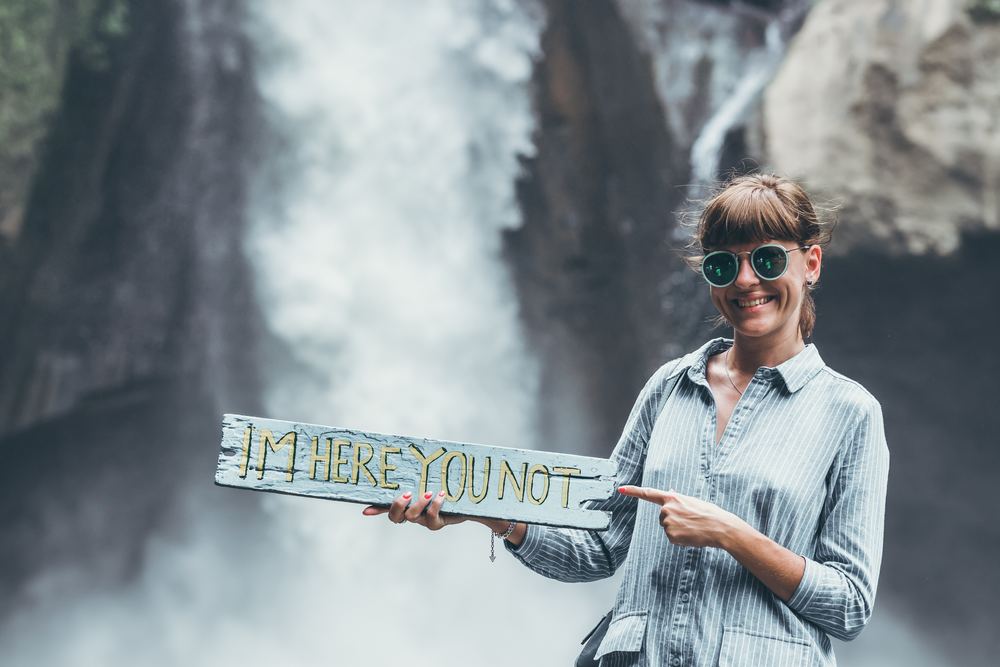 girl in front of waterfall holding board meant to cause envy that reads "I'm here, you not"